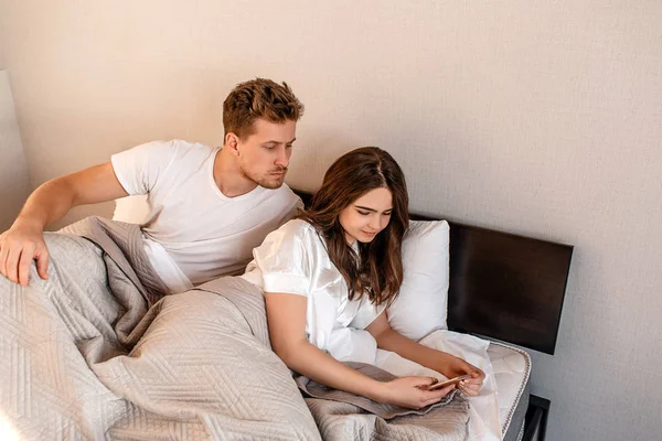 Casal jovem na cama. Homem sério está espionando suas namoradas telefone, problemas de relacionamento — Fotografia de Stock