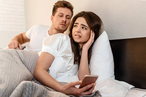 Junges Paar im Bett. schöne Frau ist nervös, während ihr Freund ihr Telefon ausspioniert, Angst vor Enthüllung — Stockfoto