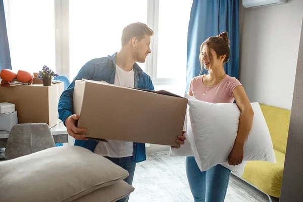 Casal jovem família comprou ou alugou seu primeiro pequeno apartamento. Eles seguram caixa e travesseiros. As pessoas olham umas para as outras e sorriem. Feliz animado . — Fotografia de Stock