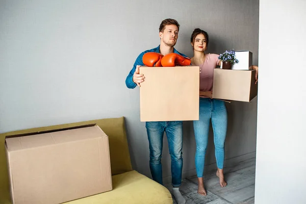 Ung familj par köpt eller hyrt sin första lilla lägenhet. De står i rummet och hålla lådor med grejer. Boxningshandskar och blommor på toppen. Allvarliga. — Stockfoto