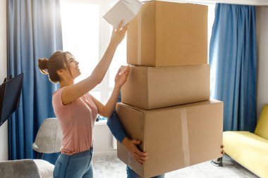 Young family couple bought or rented their first small apartment. Guy hold three cardboards. Young woman look up and reach box on top. Unpacking. clipart