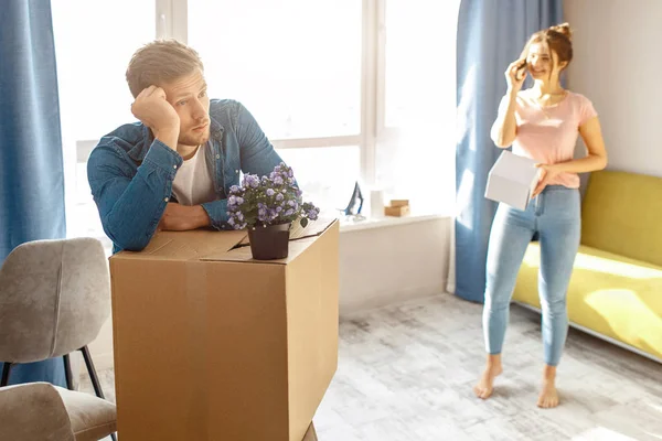 Young family couple bought or rented their first small apartment. Bored man lean on box. Young woman talk on phone. People unpacking after moving into apartment.
