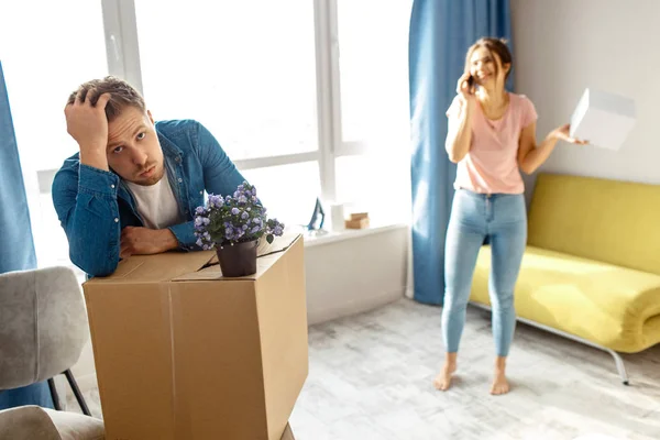 Young family couple bought or rented their first small apartment. Tired bored man looks exhausted. Young woman stand and talk on phone. Moving in and unpacking.