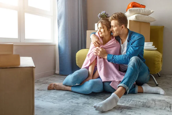 Mladý pár rodinných koupil nebo si pronajal jejich první malý byt. Muž a žena spolu sedí a obejmout. Ramena zakrýt růžovou dekou. — Stock fotografie
