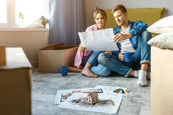 Mladý pár rodinných koupil nebo si pronajal jejich první malý byt. Sedět na podlaze a podívat se společně na byt plán. Nastěhování a rozbalení. — Stock fotografie