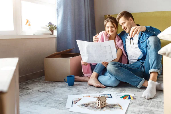 Mladý pár rodinných koupil nebo si pronajal jejich první malý byt. Veselí lidé spolu sedět na podlaze a podívejte se na plán bytu. Chlap drží klíče. Pozorný a šťastná. — Stock fotografie