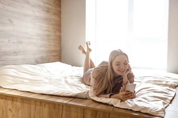 Young beautiful blonde woman lying on bed this morning. She look at phone in hands and smile. Bright daylight comes out of window. Relaxed and peaceful.