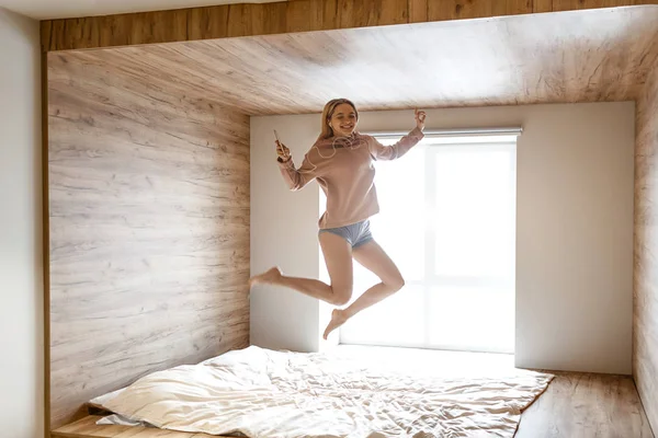 Young beautiful blonde very happy woman jump up on bed this morning. She smile and enjoy. Model listen to music through earphones. Daylight comes out of window. — Stock Photo, Image