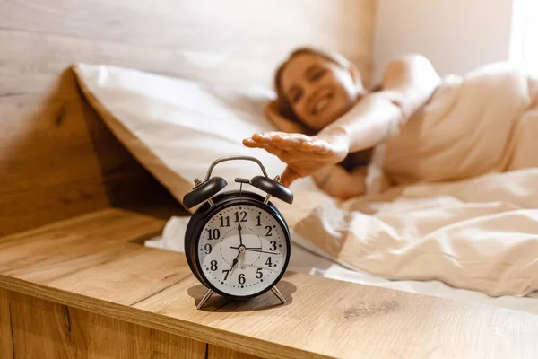 Νέοι όμορφη ξανθιά γυναίκα ξαπλωμένη στο κρεβάτι το πρωί. Θετικό πρότυπο ευτυχισμένη φθάνοντας το ρολόι συναγερμών με το χέρι. . Φως της ημέρας. Καλή διάθεση. — Φωτογραφία Αρχείου