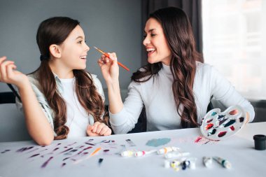 Güzel esmer beyaz anne ve kızı birlikte odaya boya. Oynak genç kadına dokun kızlar burun fırça ve gülüyor. Anne Çocuk bakmak. Onlar birlikte masaya oturup.