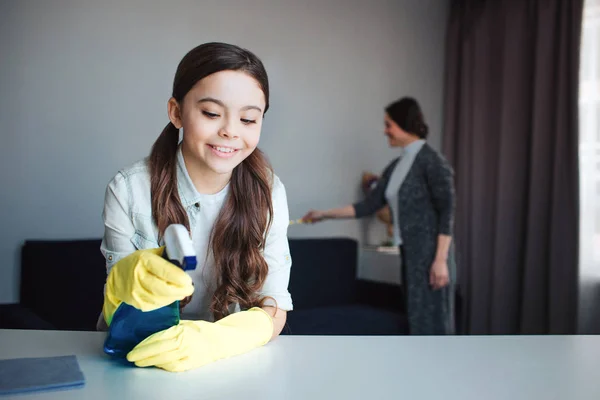 Gyönyörű barna kaukázusi anya és lánya együtt takarítás a szobában. Lány elé állni, és használja a permetező-ra táblázat. Sárga kesztyűt visel. Neki anya tiszta mögött. — Stock Fotó