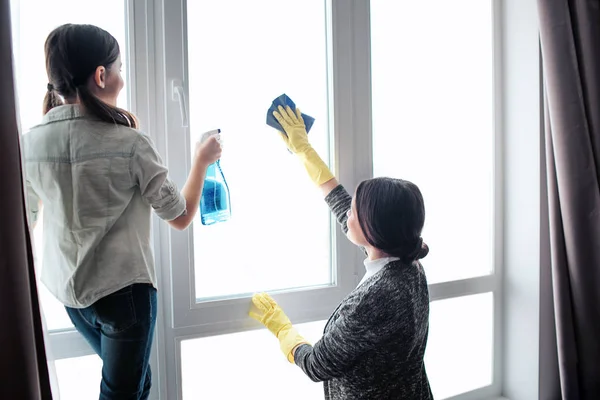 Linda morena caucasiana mãe e filha janela de limpeza juntos no quarto. Spray de uso feminino. Jovem mulher lavando janela. Trabalham juntos. . — Fotografia de Stock