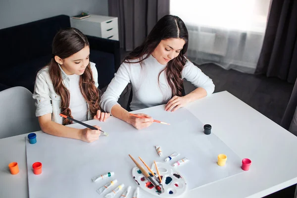 Belle brune caucasienne mère et fille peignent ensemble dans la chambre. Ils travaillent à la même table sur un bout de papier blanc. Calme et concentré . — Photo