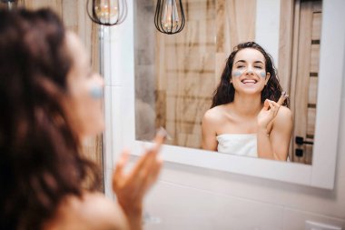 Sabah akşam rutin ayna adlı genç sportif esmer güzel kadının yapması. Neşeli mutlu modeli göz ayna ve gülümseme. O yüz ve burun üzerinde bazı krem koymak.