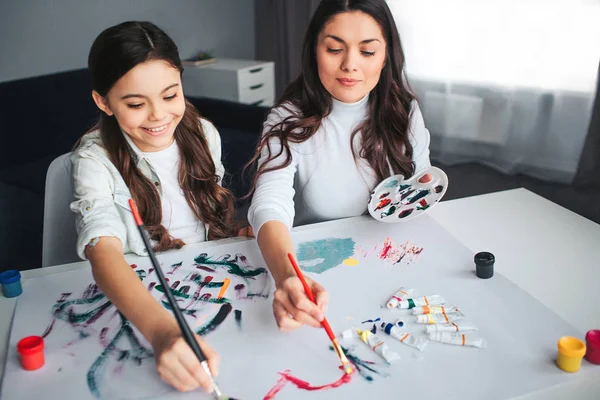 Beautiful brunette caucasian mother and daughter paint together in room. Happy girl hold brushes with mom. She smile. Young woman has palette in hand. Painting heart shape.