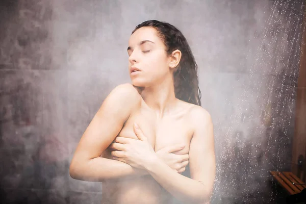 샤워에 젊은 매력적인 섹시 한 여자. 자신을 세척 하는 동안의 즐거움 손으로 가슴을 덮고. 눈 폐쇄. 유리 벽에는 물 증기 세미 흐린된 사진. — 스톡 사진