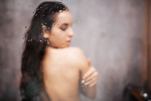 Young attractive sexy woman in shower. Standing back to camera. Embrace herself with closed eyes. Blurred photo. Water vapor on glass wall. — Stock Photo, Image