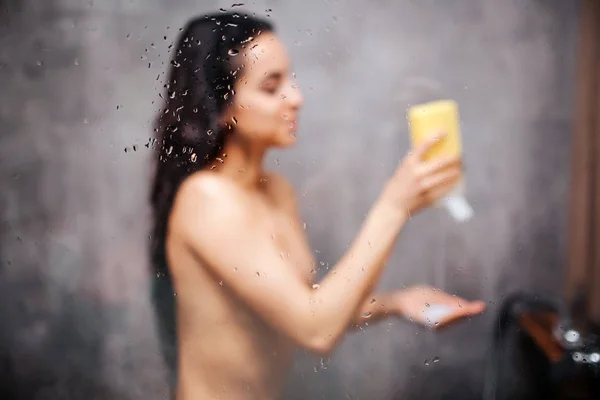 Joven atractiva mujer sexy en la ducha. Precioso modelo de pelo oscuro poner un poco de gel de ducha en la mano y sonreír. Está bajo la corriente del agua. Imagen borrosa. Vapor de agua en pared de vidrio . —  Fotos de Stock