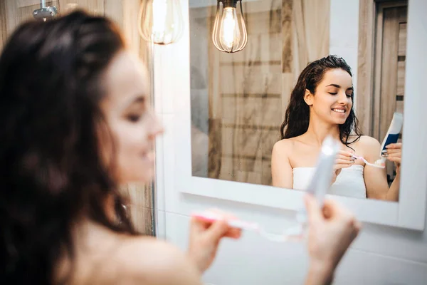 Jeune femme belle aux cheveux bruns et sportive faisant la routine du matin au miroir. Elle portait une serviette et souriait. Modèle tenir tube à crème dans les mains . — Photo