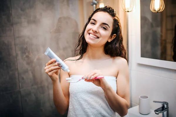 Jeune femme belle aux cheveux bruns et sportive faisant la routine du matin au miroir. Elle regarde la caméra et sourit. Mettre de la pâte à dents sur la brosse à dents . — Photo