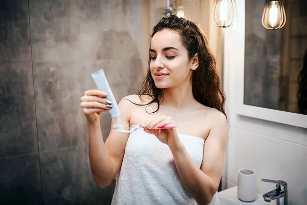 Jeune femme belle aux cheveux bruns et sportive faisant la routine du matin au miroir. Elle a mis de la pâte dentaire sur une brosse à dents. Beau modèle regardez-le. Corps enveloppé avec une serviette blanche . — Photo