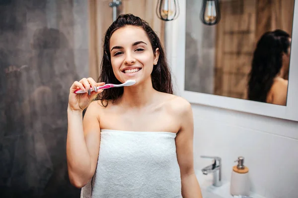 Jeune femme belle aux cheveux bruns et sportive faisant la routine du matin au miroir. Elle regarde la caméra et sourit. Modèle tenir la pâte dentaire avec brosse à dents près de la bouche. Poser sur la caméra . — Photo