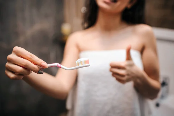 Jeune femme belle aux cheveux bruns et sportive faisant la routine du matin au miroir. Vue découpée du modèle en serviette blanche tenant brosse à dents avec pâte à dents et gros pouce vers le haut . — Photo