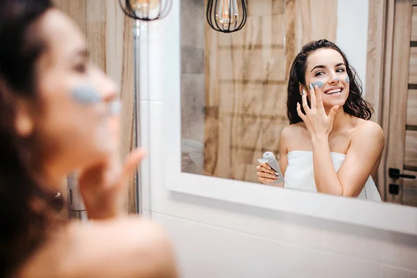 Young sporty dark-haired beautiful woman doing morning evening routine at mirror. Cheerful happy model wrapped in white towel and put mask on face.