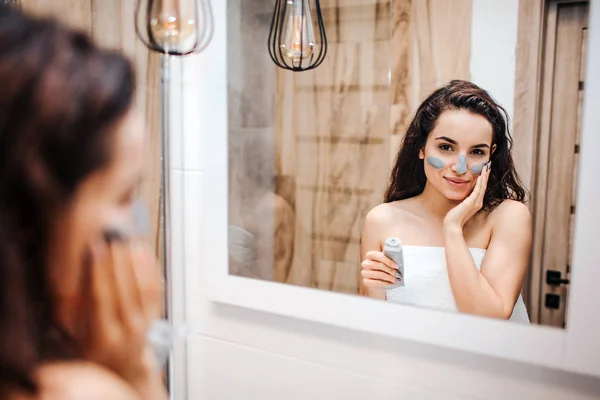 Jeune femme belle aux cheveux bruns et sportive faisant la routine du matin au miroir. Elle se regarde dans le miroir et met un masque sur le visage . — Photo