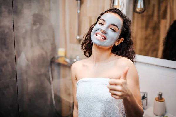 Jeune femme belle aux cheveux bruns et sportive faisant la routine du matin au miroir. Joyeux regard heureux modèle sur la caméra et msile. Elle a un masque. Femme tenir gros pouce vers le haut . — Photo