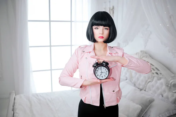 Fashion freak. Glamour synthetic girl, fake doll with empty look and short black hair is holding clock while standing in the bed. Stylish woman in pink jacket on the bedroom. Fashion and beauty