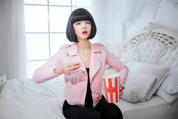 Fashion freak. Glamour synthetic interested girl, fake doll with short black hair is holding popcorn and looking TV while sitting in the bed. Stylish woman in pink jacket on the bedroom. Fashion and