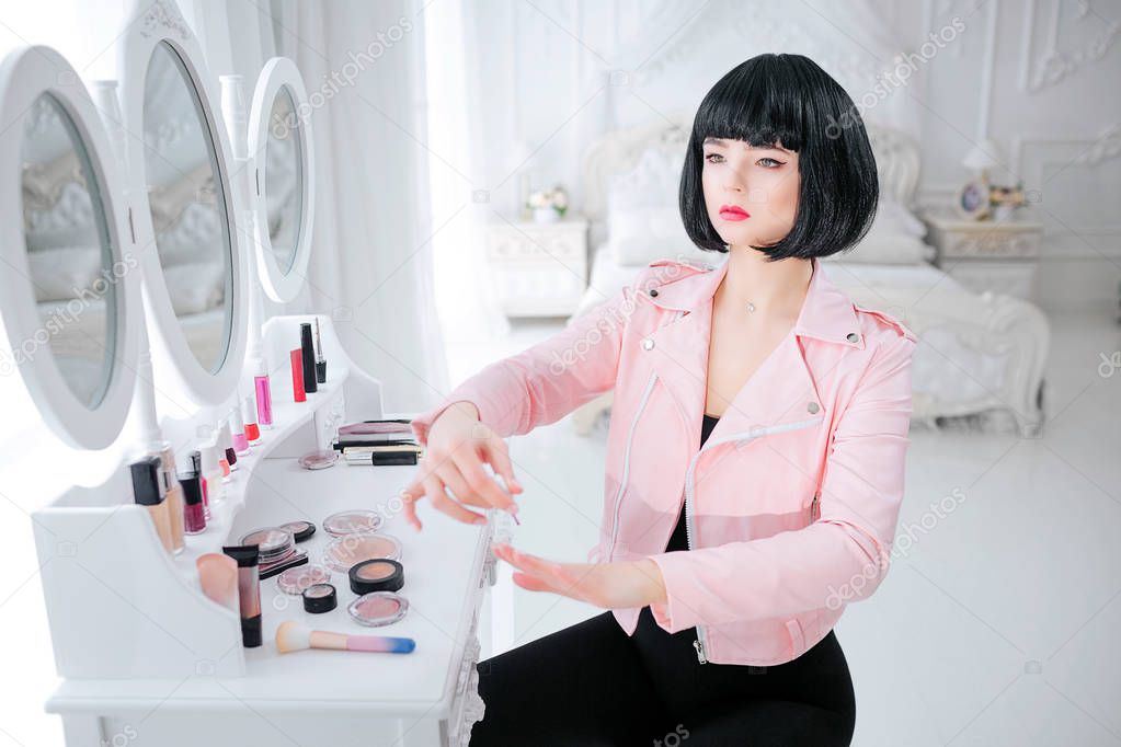 Fashion freak. Glamour synthetic girl, fake doll with empty look and short black hair is painting her nails while sitting near mirror. Stylish woman in pink jacket in the bedroom. Fashion and beauty