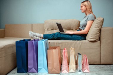 Evde online alışveriş. Genç, gülümseyen bir kadın koltukta oturuyor ve renkli alışveriş torbalarının yanında dizüstü bilgisayar kullanıyor.