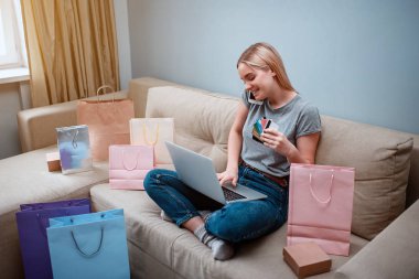 Evde alışveriş online. Genç gülümseyen alışveriş dizüstü bilgisayar ve kredi kartları ile alışveriş torbaları ile bir kanepede otururken smarthphone online shop tarafından emir veriyor