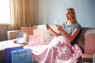 Evde online alışveriş. Tabletli ve akıllı telefonlu genç sarışın kadın, kanepede battaniyeyle otururken online dükkandaki malların fiyatlarını karşılaştırıyor.