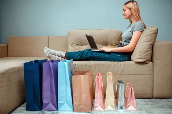 Compras en línea en casa. Joven mujer sonriente está sentado en el sofá y el uso de ordenador portátil cerca de coloridas bolsas de compras — Foto de Stock