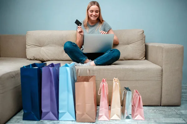 Compras en línea en casa. Joven mujer feliz con tarjeta de crédito está listo para grandes ventas en la tienda en línea mientras está sentado en el sofá con bolsas de compras — Foto de Stock