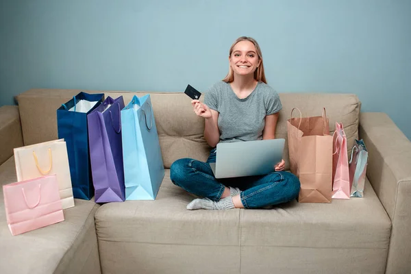 Compras en línea en casa. Joven mujer feliz con tarjeta de crédito está listo para el día de solteros en un sofá cerca de las bolsas de compras — Foto de Stock