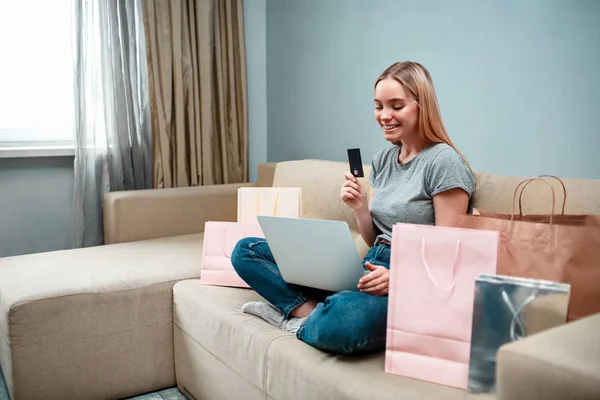 Compras en línea en casa. Joven mujer sorprendida con tarjeta de crédito está pidiendo en la tienda en línea mientras está sentado en un sofá con bolsas de compras — Foto de Stock