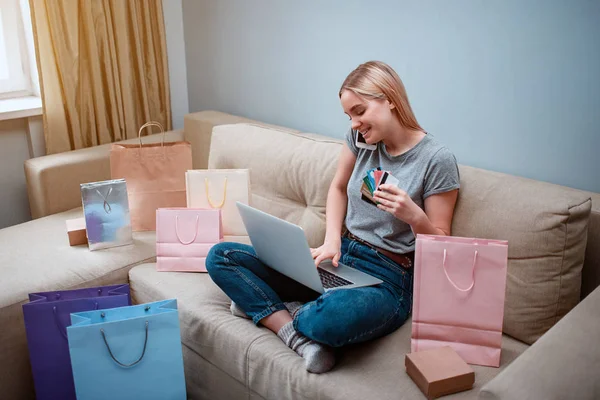 Compras en línea en casa. Joven comprador sonriente con computadora portátil y tarjetas de crédito está ordenando por teléfono inteligente en la tienda en línea mientras está sentado en un sofá con bolsas de compras — Foto de Stock