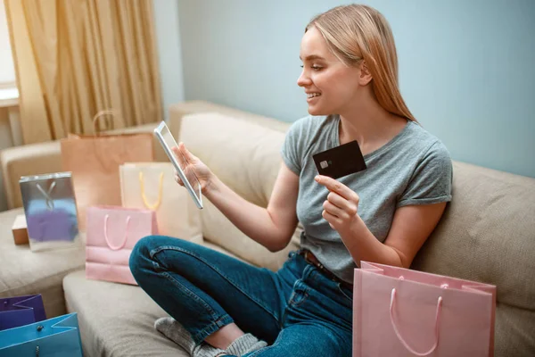 Compras en línea en casa. Joven comprador feliz con tarjeta de crédito y tableta está comprando en la tienda en línea mientras está sentado en un sofá con bolsas de compras — Foto de Stock