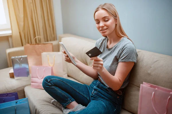 Compras en línea en casa. Joven comprador feliz con tarjeta de crédito y tableta está comprando en la tienda en línea mientras está sentado en un sofá con bolsas de compras — Foto de Stock