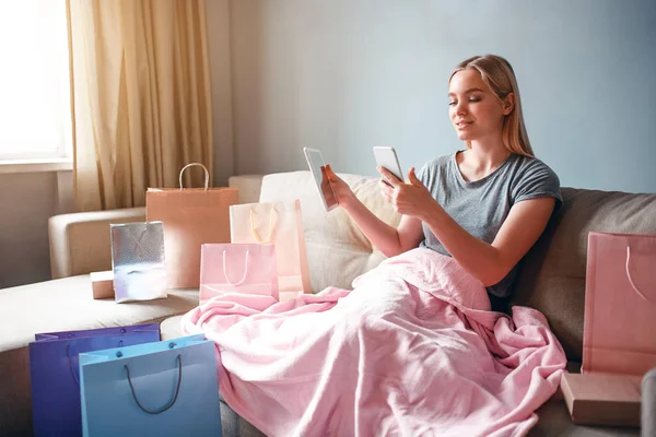 Compras en línea en casa. Mujer rubia joven con tableta y teléfono inteligente está comparando los precios de los bienes en la tienda en línea mientras está sentado con manta en un sofá — Foto de Stock