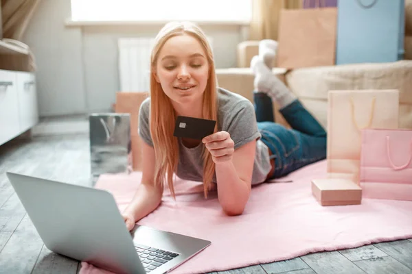 Online shopping at home. Young happy woman ia ready to spend money from husbands bank card on black Friday while lying on the floor with shopping bags