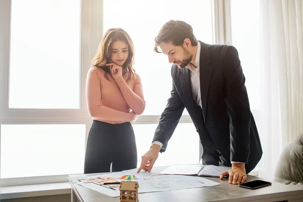 Couple buy or rent apartment together. They stand upon table and hold there plan. Young man look at it. He lean to table. Young woman look down too and pose. Daylight.