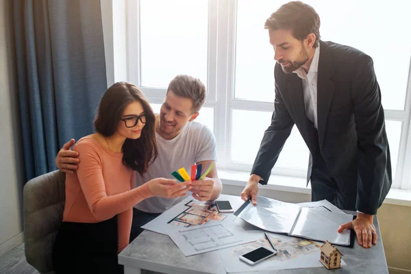 Couple buy or rent apartment together. They sit in front of realtor. Young woman look at colorful tissue pieces in hand and smile. Man embrace her and look at woman.