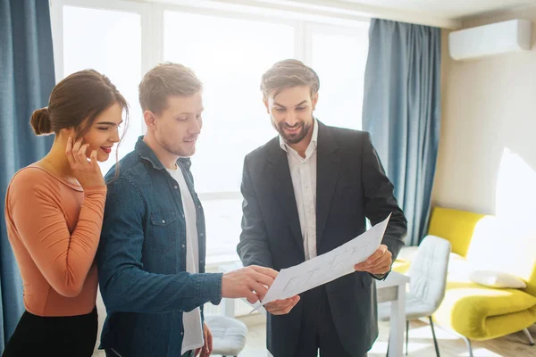 Couple buy or rent apartment together. Realtor and man hold plan together and look at it. Woman stand behind guy. Business deal. Happy and positive.