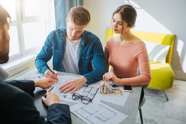 Pareja comprar o alquilar apartamento juntos. Un hombre serio y concentrado puso la firma en los documentos. Trato de negocios. Jovencita, míralo. Realtor sentarse frente a ellos . — Foto de Stock