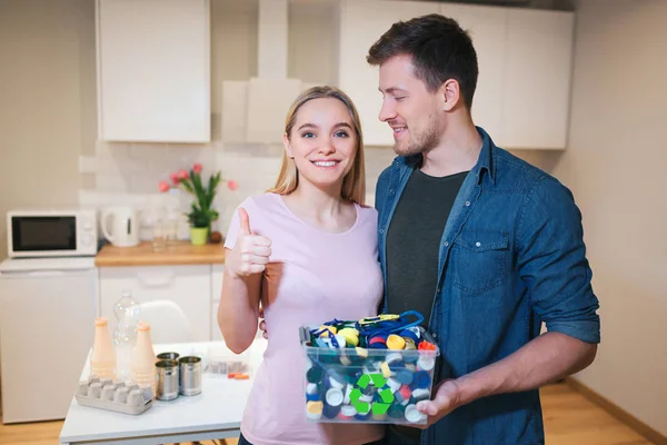 Proteger o ambiente. Jovem família sorrindo segurando recipiente de reciclagem preenchido com resíduos eletrônicos no fundo da cozinha — Fotografia de Stock